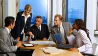 A group of five people in business attire engaged in a meeting around a conference table, with papers and a laptop present.
