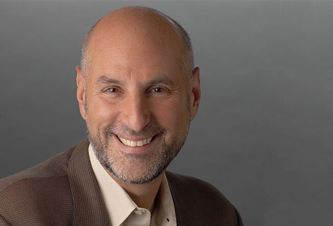 Headshot of a smiling person with a bald head and beard, wearing a brown blazer and light-colored shirt, against a gray background.