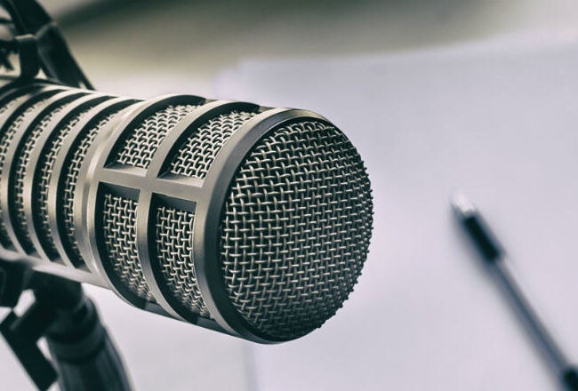 Close-up of a condenser microphone with a blurred background of white paper and a pen, often representing podcasting or broadcasting.