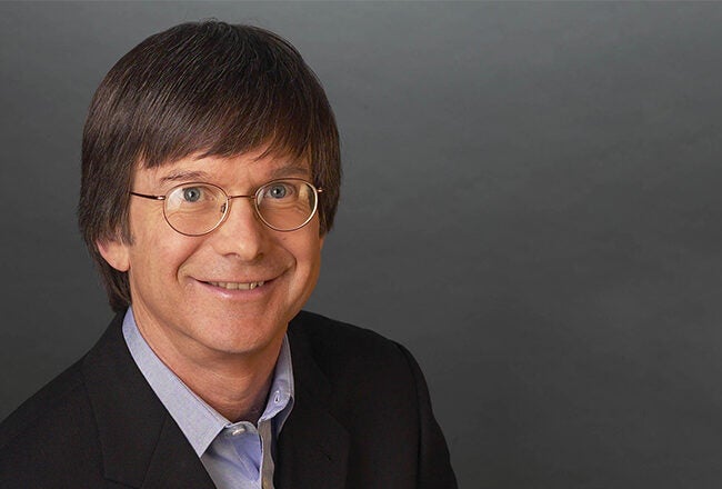 A headshot of a person wearing glasses and a suit jacket, smiling against a dark gray background.