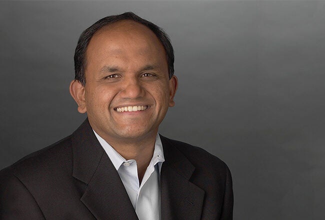 A person wearing a dark suit jacket and white shirt, smiling against a plain gray background. It''s a professional headshot.