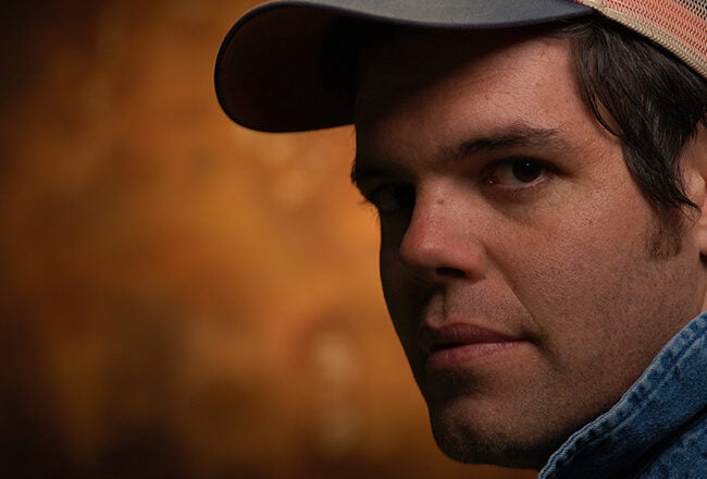 Headshot of a person wearing a cap and denim jacket, looking over their shoulder against a warm, blurred background.