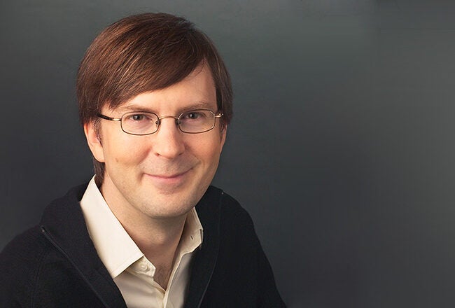 A headshot of a person with glasses, wearing a light-colored shirt and a dark sweater, smiling against a dark background.