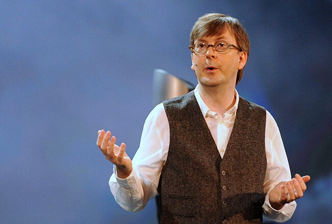 A person in a white shirt and dark vest gestures while speaking on stage, set against a blurred blue and gray background.