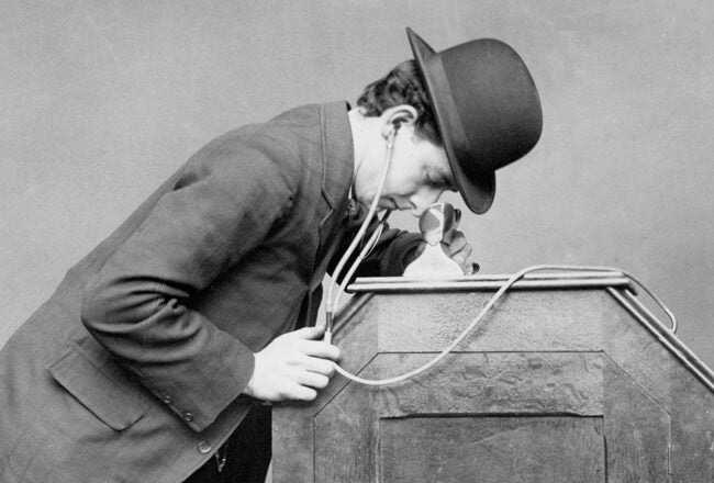 A person wearing a bowler hat leans in to look and listen through a large viewing device. The image represents early audio-visual technology.