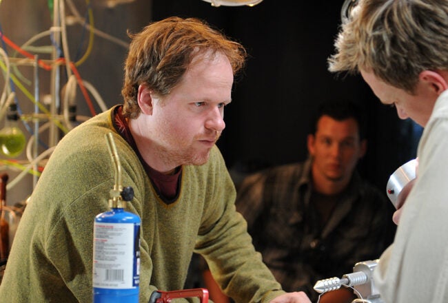 Two people are having a discussion at a workbench, surrounded by scientific equipment and colorful tubes. Another person is seated in the background, observing.