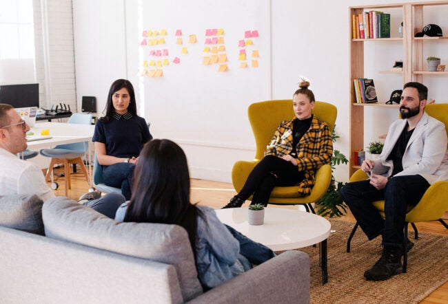 A group of people engaged in a casual meeting in a modern office space. Some are seated on sofas and armchairs, and colorful sticky notes are visible on the wall.