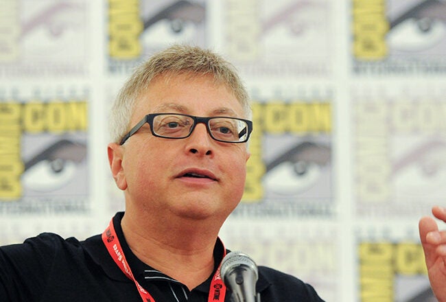 A person speaking at a microphone with hands raised, wearing glasses and a red lanyard, against a backdrop with Comic-Con logos.