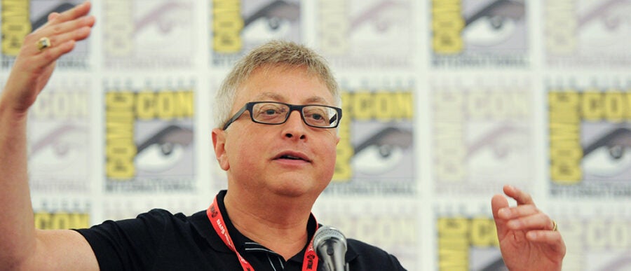 A person speaking at a microphone with hands raised, wearing glasses and a red lanyard, against a backdrop with Comic-Con logos.