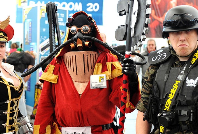 Three people dressed in elaborate cosplay costumes. The first wears a military-style outfit with a red hat and holds a futuristic weapon. The second is in a red robe with a mask and carries a large prop weapon. The third wears a tactical vest, cam