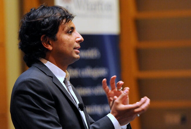 A person in a suit giving a presentation, standing profile with hands gesturing, in a conference or lecture setting.