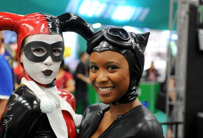 Two people dressed in costumes; one in a Harley Quinn outfit with red and black jester attire, and the other in a black Catwoman suit, smiling at a convention.