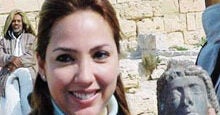 A person smiling while holding a small stone sculpture of a human head, with a stone wall and others in the background.