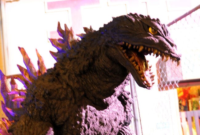 A large sculpture of a monster resembling Godzilla with sharp teeth and dorsal fins is displayed in a brightly lit setting.