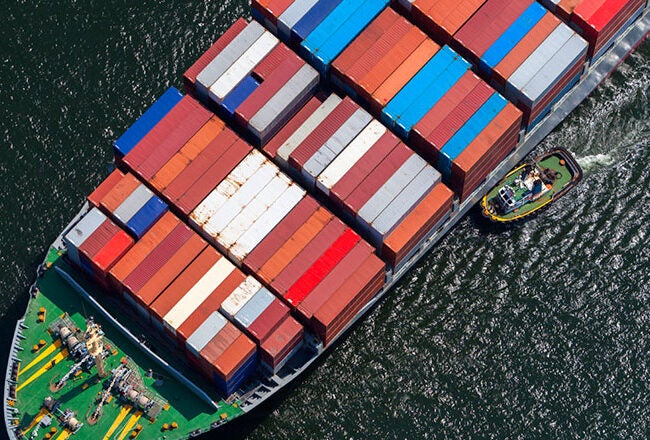 An aerial view of a large cargo ship loaded with colorful shipping containers, accompanied by a smaller tugboat on the water.