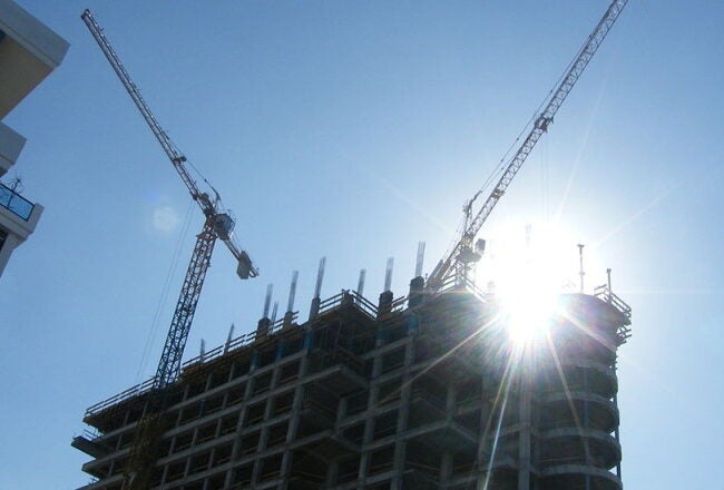 A building under construction with cranes against a clear blue sky, with the sun shining brightly behind the structure.