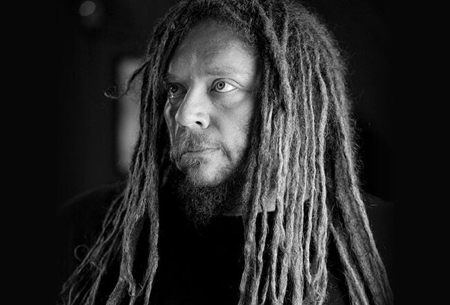 A black-and-white headshot of a person with long dreadlocks and a beard, looking slightly to the side.