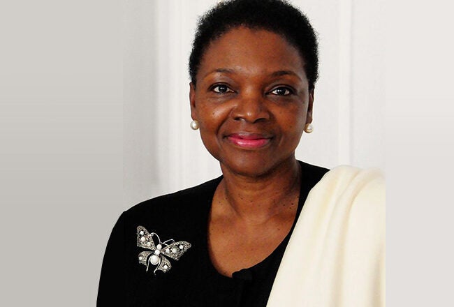Headshot of a person wearing a black outfit with a butterfly brooch and a white scarf, against a light background.