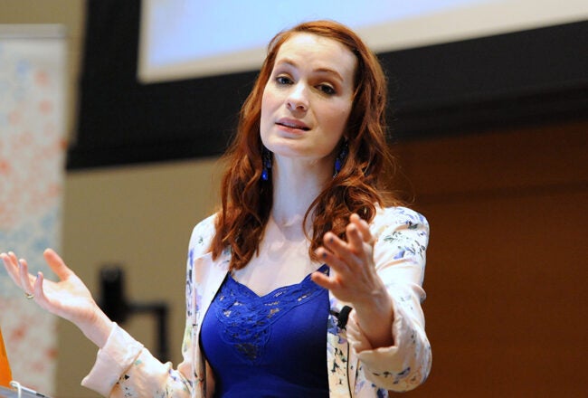 A person speaking passionately at a podium, with an orange laptop. They are indoors, possibly at a conference or presentation.