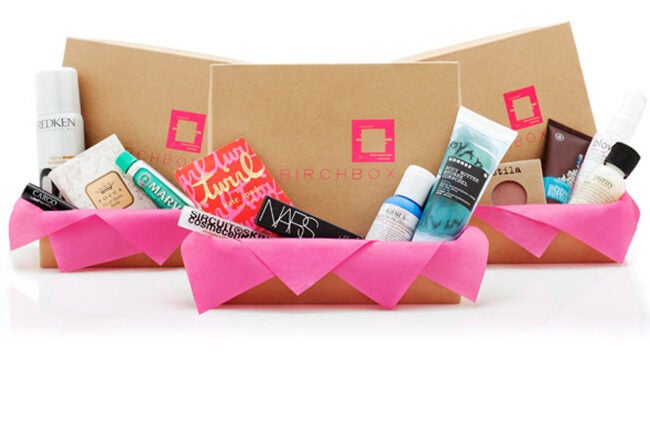 Gift boxes with skincare and beauty products, surrounded by pink tissue paper. Includes items such as moisturizers, sprays, and lotions.