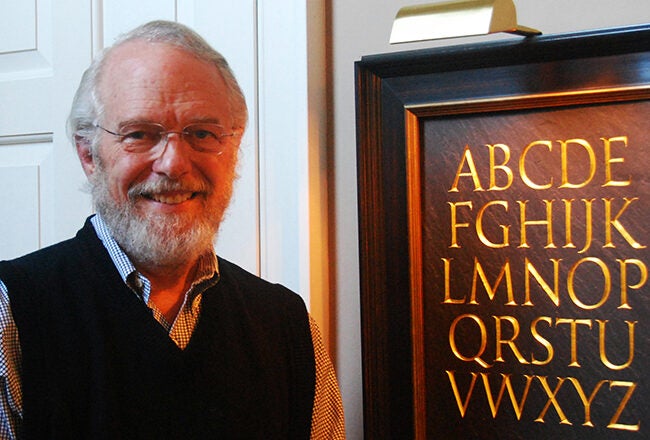 A person smiling next to a framed alphabet display with gold lettering.
