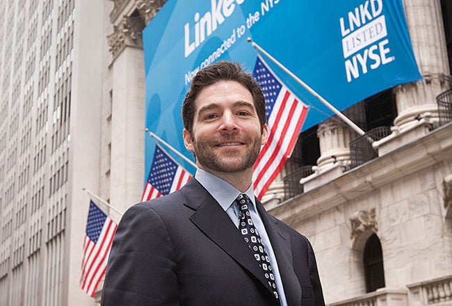 A person in formal attire stands in front of the New York Stock Exchange building adorned with American flags and a LinkedIn banner announcing its listing.