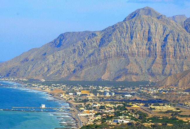 A coastal town with buildings nestled along a shoreline, next to a vast blue sea. Majestic rocky mountains rise in the background.