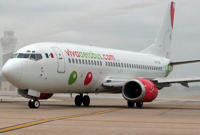 An airplane from VivaAerobus taxiing on an airport runway. The aircraft is predominantly white with red and green accents, featuring the airline''s logo on the fuselage.