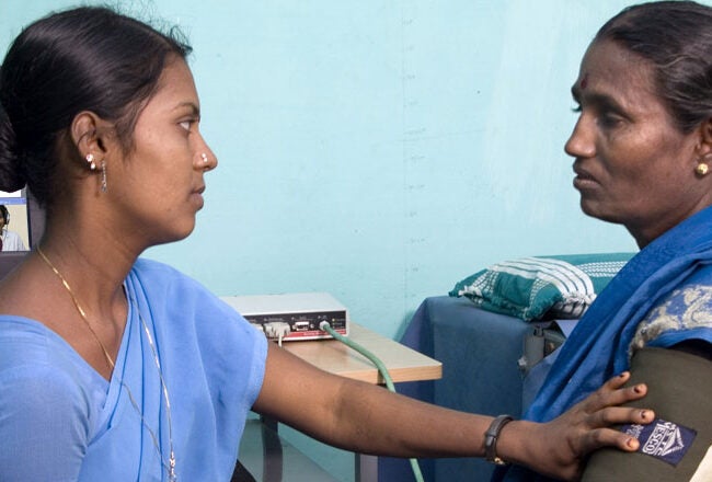 A healthcare worker is taking a patient''s blood pressure in a clinical setting. A computer is in the background displaying medical software.