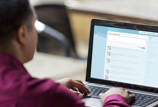 A person in a red shirt using a laptop with a social media platform, likely Twitter, displayed on the screen.