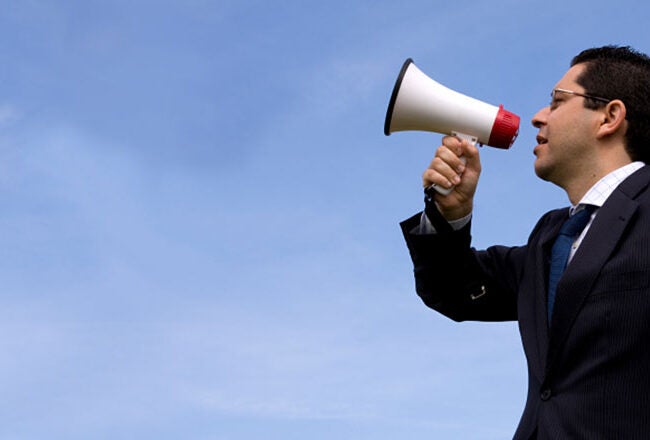 A person in a suit using a megaphone against a clear blue sky, representing communication or public speaking.