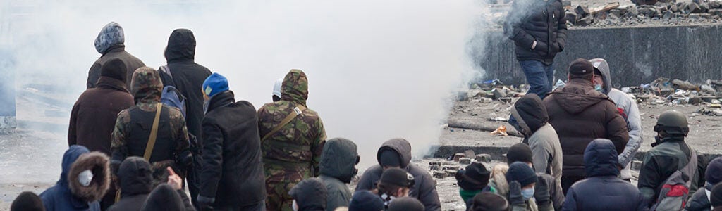 A group of people in heavy clothing gather in a street surrounded by smoke and debris, possibly during a protest or conflict.