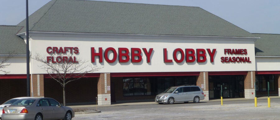 The image shows the exterior of a Hobby Lobby store with parking in front. The store''s signage includes "Crafts," "Floral," "Frames," and "Seasonal."