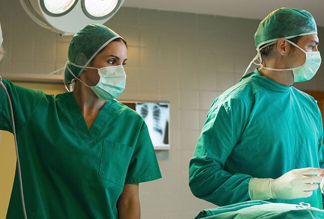 Medical professionals in surgical attire, including masks and caps, are in an operating room, preparing for or conducting surgery. Equipment and an operating table are visible.