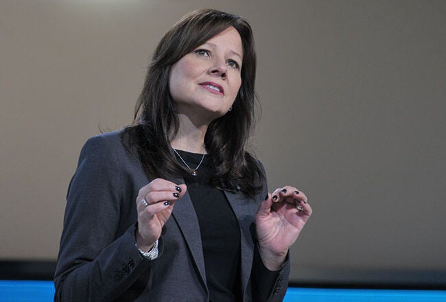 An individual in a blazer speaking on stage, gesturing with their hands, possibly during a presentation or speech.