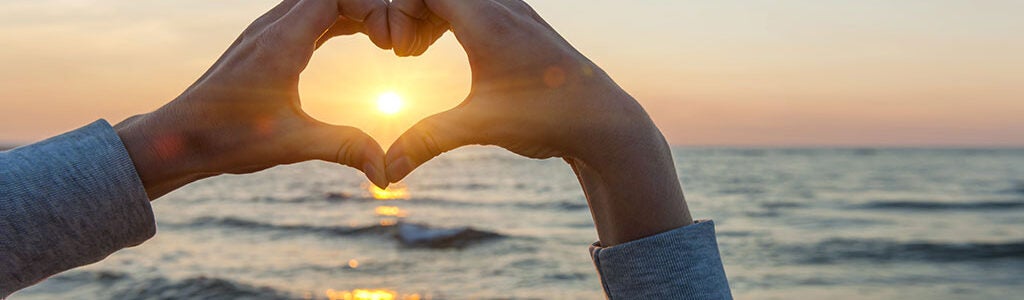 Hands forming a heart shape with the sun in the center, set against a backdrop of the ocean and a sunset.