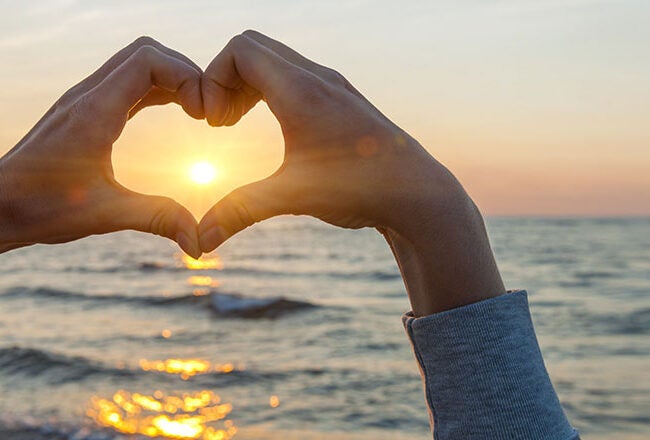 Hands forming a heart shape with the sun in the center, set against a backdrop of the ocean and a sunset.