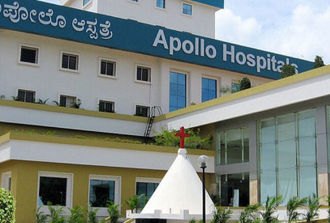 A multi-story building with a sign reading "Apollo Hospitals" in English and another language. The structure features white walls, large windows, and greenery in the foreground.