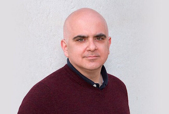 A headshot of a person wearing a maroon sweater over a black shirt, with a neutral expression, against a light-colored background.