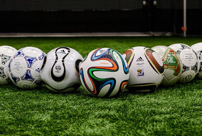 A row of various Adidas soccer balls displayed on a grassy field, showcasing different designs and patterns over the years.