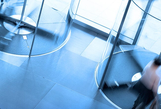 Blurred image of people moving through modern revolving glass doors in a building entrance, suggesting motion and activity.