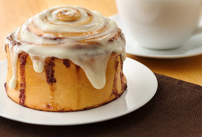 A cinnamon roll with icing on a white plate, placed on a brown napkin, and a white cup in the background on a light wood table.