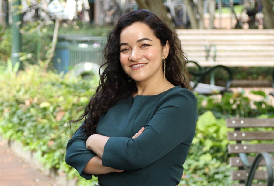 A person with long curly hair is smiling with arms crossed, standing in a park with greenery and benches in the background.