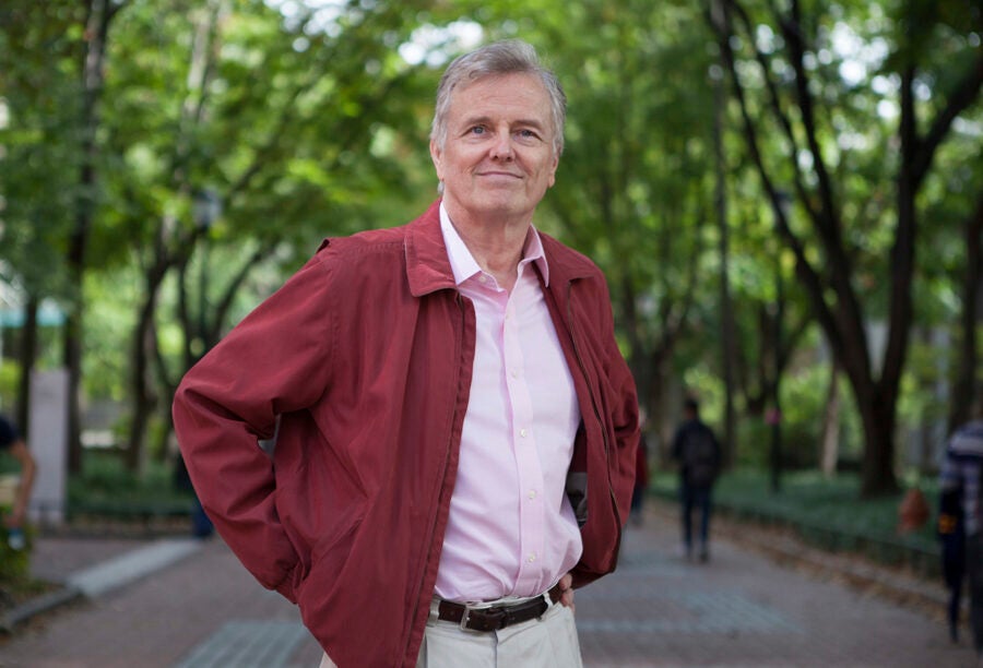 A person wearing a red jacket and pink shirt poses with hands on hips in a tree-lined park setting.