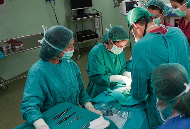 Medical team performing surgery in an operating room, wearing green scrubs, masks, and gloves, with surgical instruments on a nearby table.