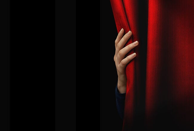A hand partially draws back a red curtain, revealing a glimpse behind it, suggesting anticipation or curiosity.