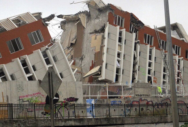 A collapsed multi-story building, tilted on its side with significant structural damage, possibly due to an earthquake. People and graffiti are visible in the foreground.