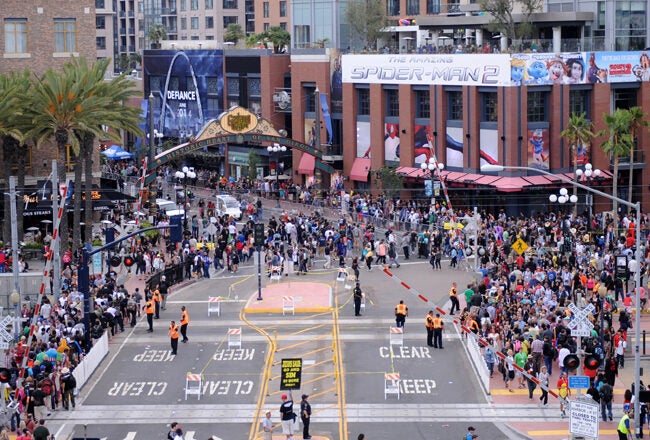 Aerial view of a bustling city street during an event, with large crowds, a Spider-Man 2 advertisement, palm trees, and a gated area.