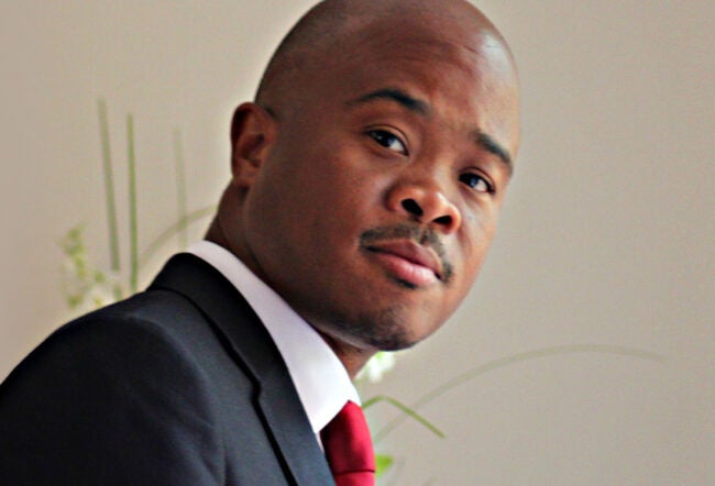 A headshot of a person wearing a dark suit, white shirt, and red tie, looking toward the camera with a neutral expression.