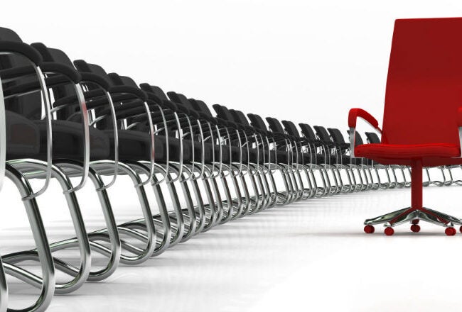 A single red office chair stands out prominently among a long row of identical black chairs, symbolizing individuality or leadership.